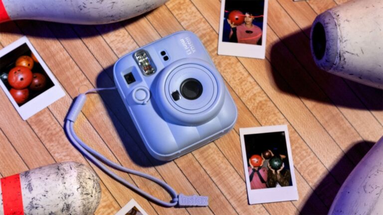 Fujifilm Instax Mini 13 instant camera, in blue, laid on the floor of a bowling alley, surrounded by fallen pins and instant prints