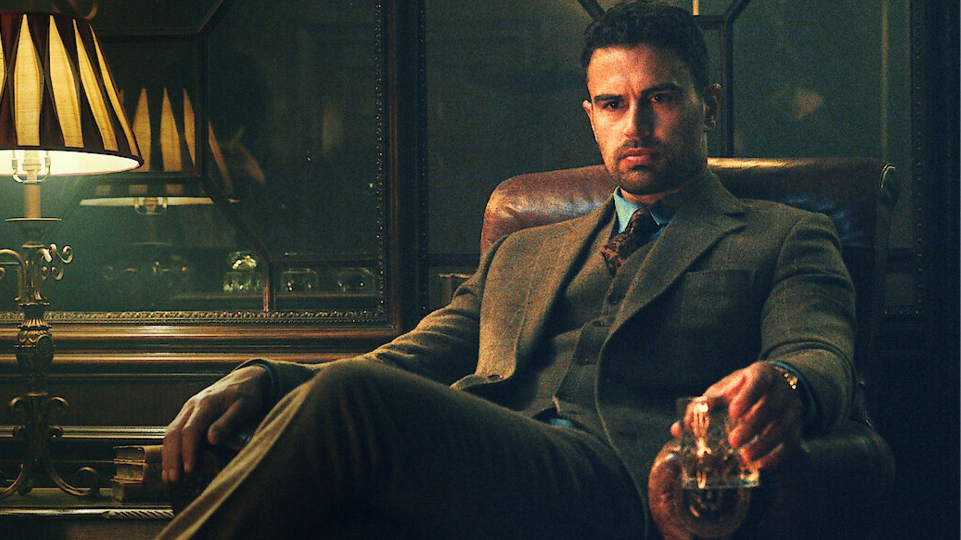 Theo James sits in a chair holding a whiskey decanter