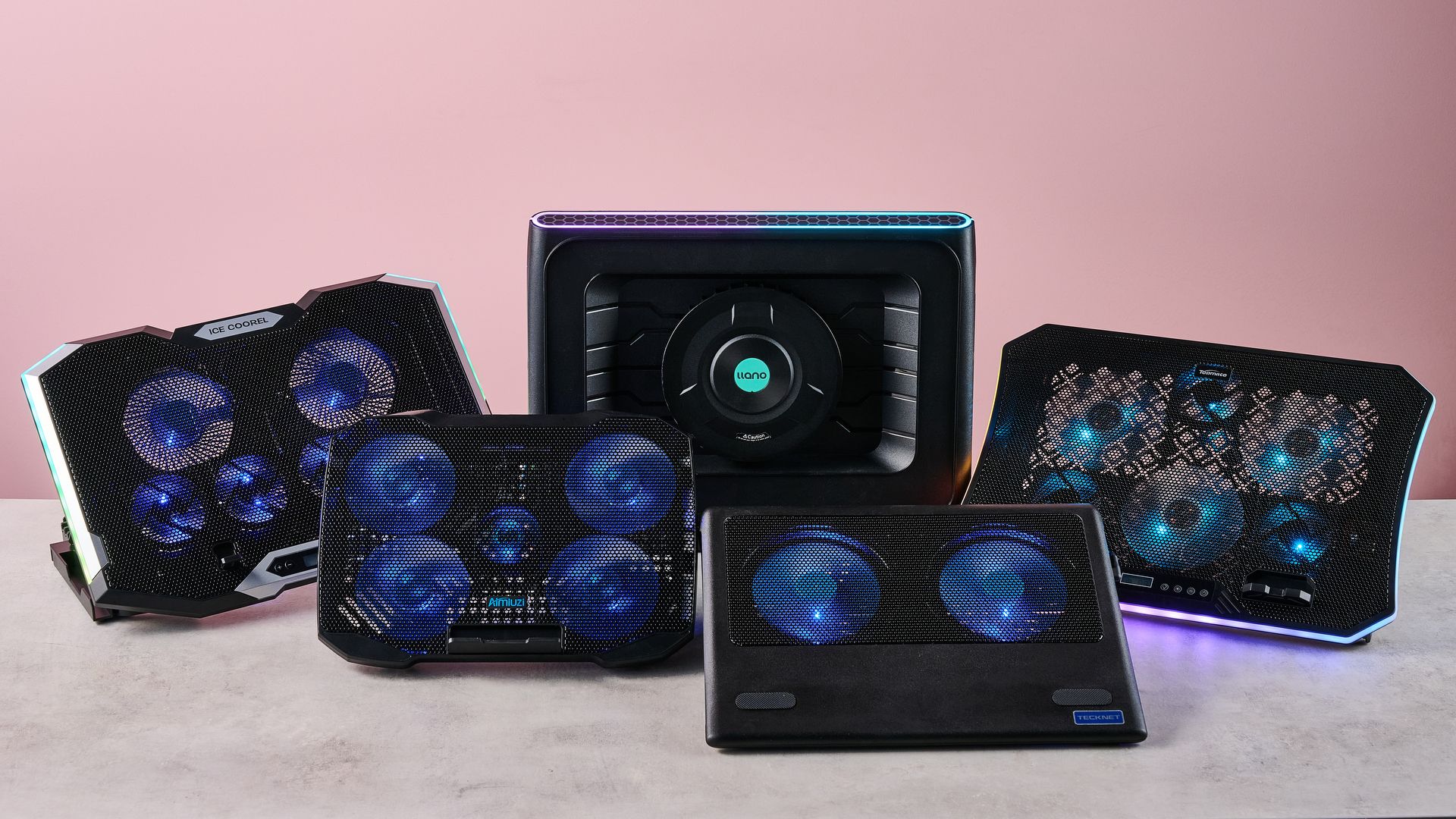 The Llano RGB Laptop Cooling Pad, TopMate C12, Aimuzi F2 Plus, ICE COOREL RGB Laptop Cooling Pad and Tecknet N8 on a gray marbled desktop in front of a pink background.