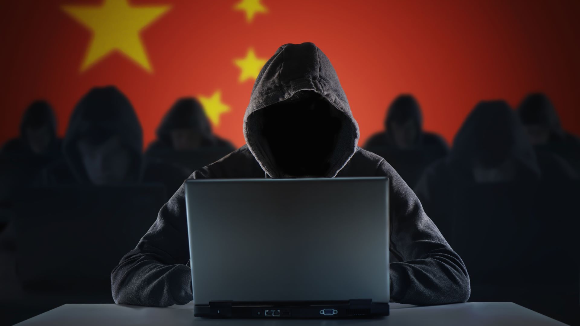 A group of hackers all wearing black with hoods pulled up over their heads with an open laptop in front of them. The background is a Chinese flag