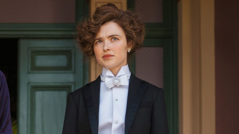Sybilla wears a suit, standing in a doorway