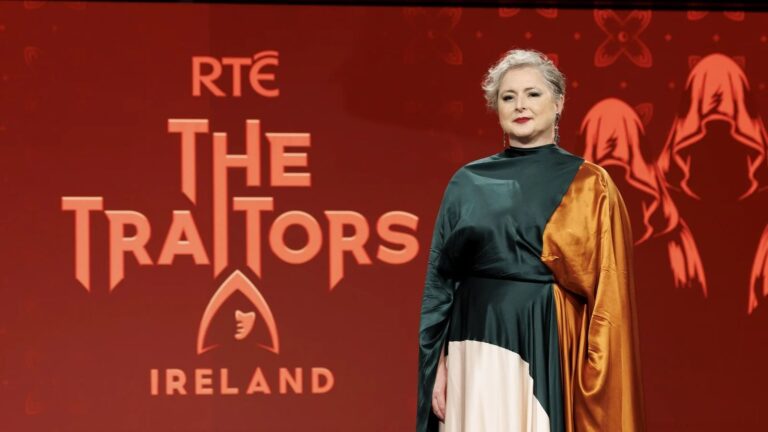 The Traitors Ireland host Siobh&aacute;n McSweeney standing in front of the logo for the show ahead of season 2.