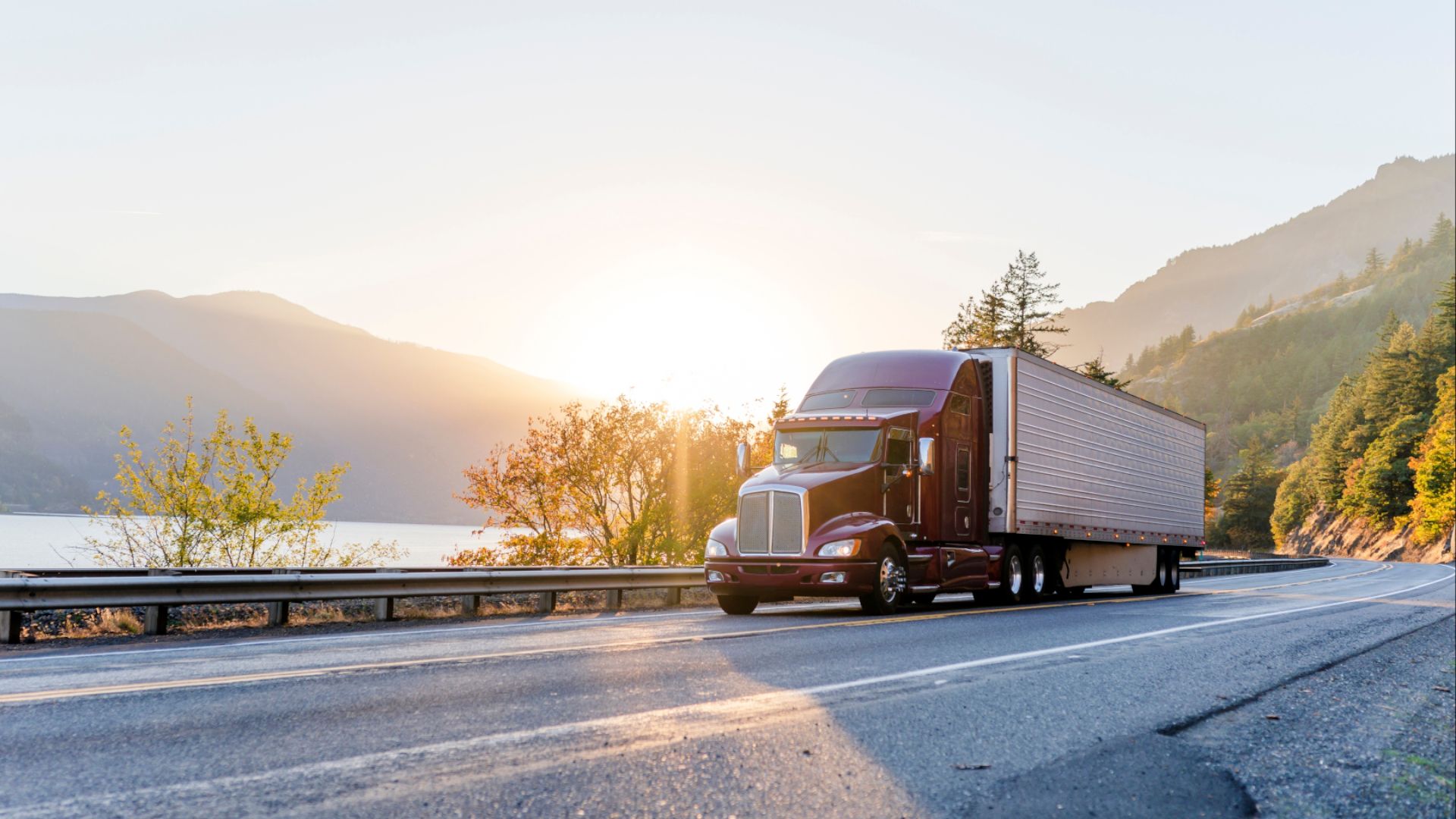 Big rig classic American powerful brown semi truck with refrigerated semi trailer transporting frozen goods running on the road along railroad and river in Columbia Gorge area with mountain ranges